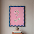 Framed artwork with a pink background and blue border on a wall above books.