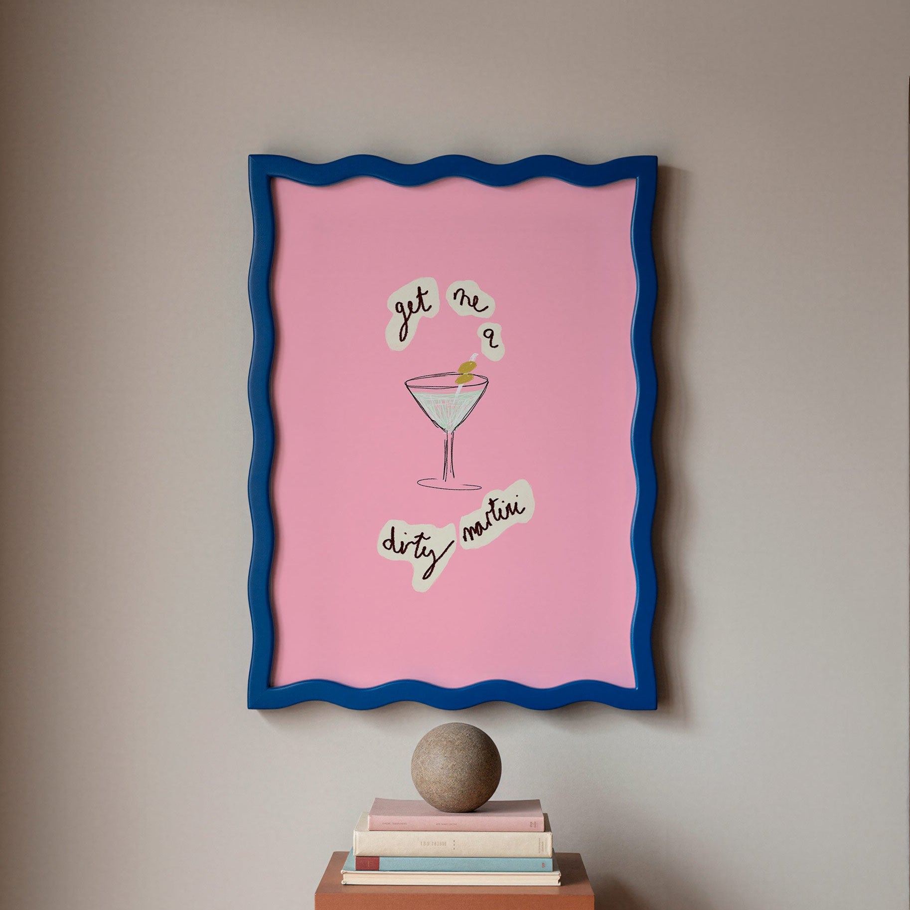 Framed artwork with a pink background and blue border on a wall above books.