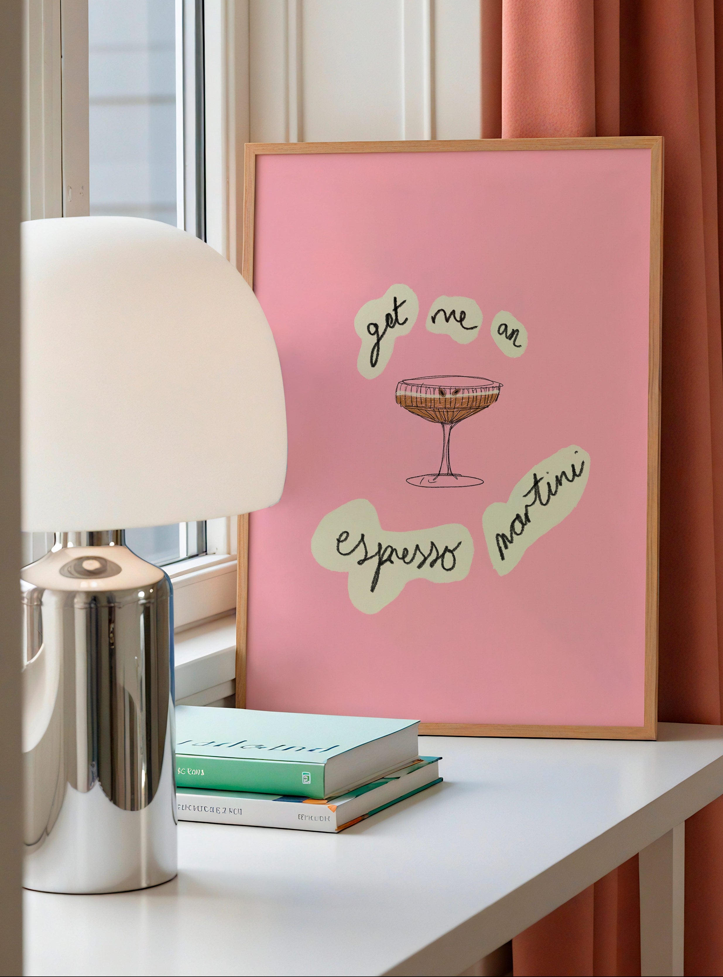 Pink framed poster with get me an espresso martini and a cocktail illustration on a white surface with books and a lamp.