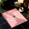 Pink tray with text on a black tiled surface with a teapot and cup.