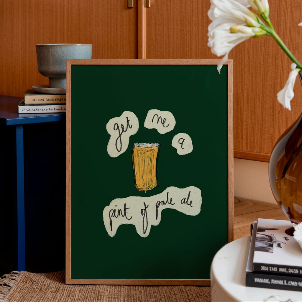 Framed artwork with text and a beer illustration on a green background, placed in a room with wooden cabinets and a vase of flowers.