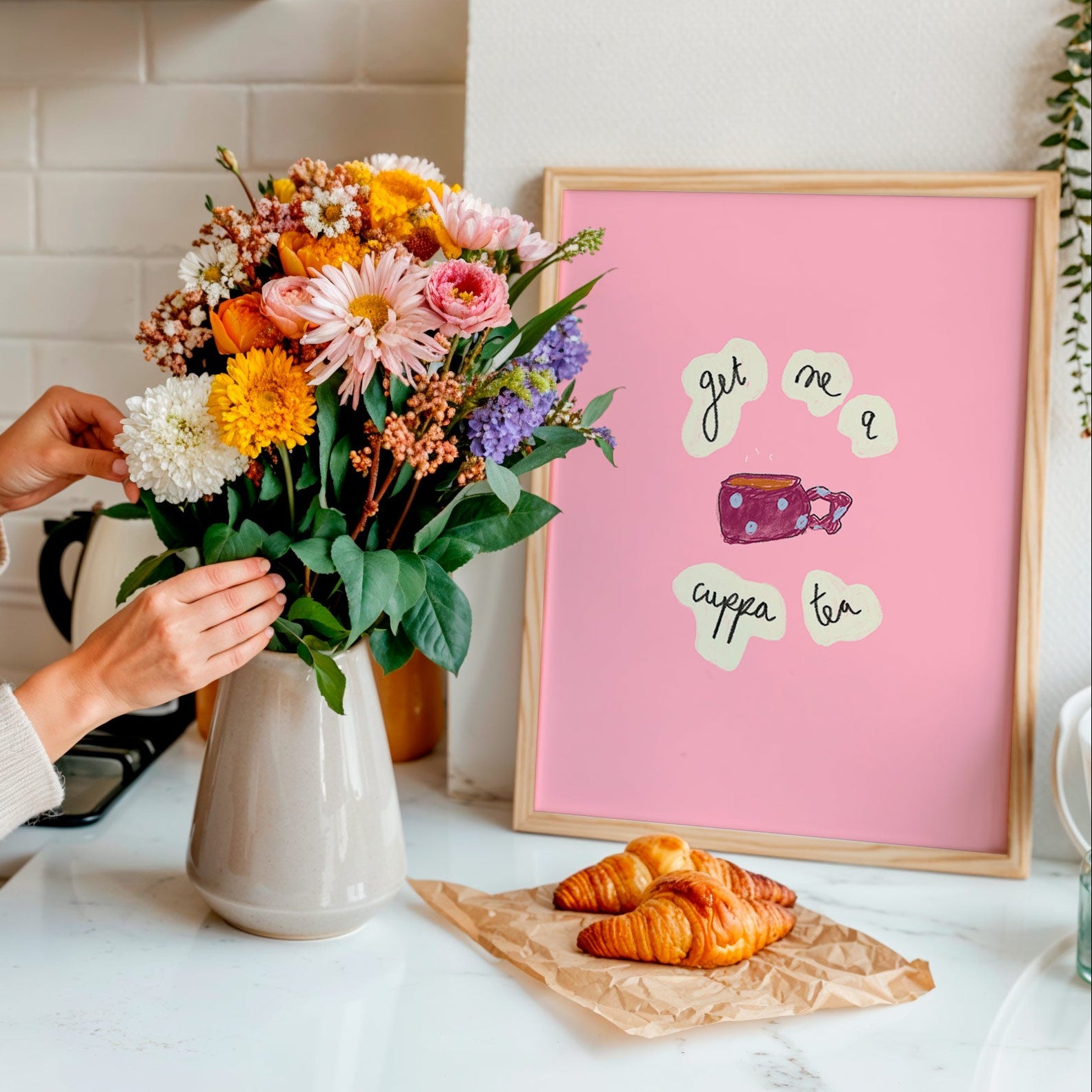 Person arranging flowers in a kitchen with a pink framed picture and pastries on the counter.