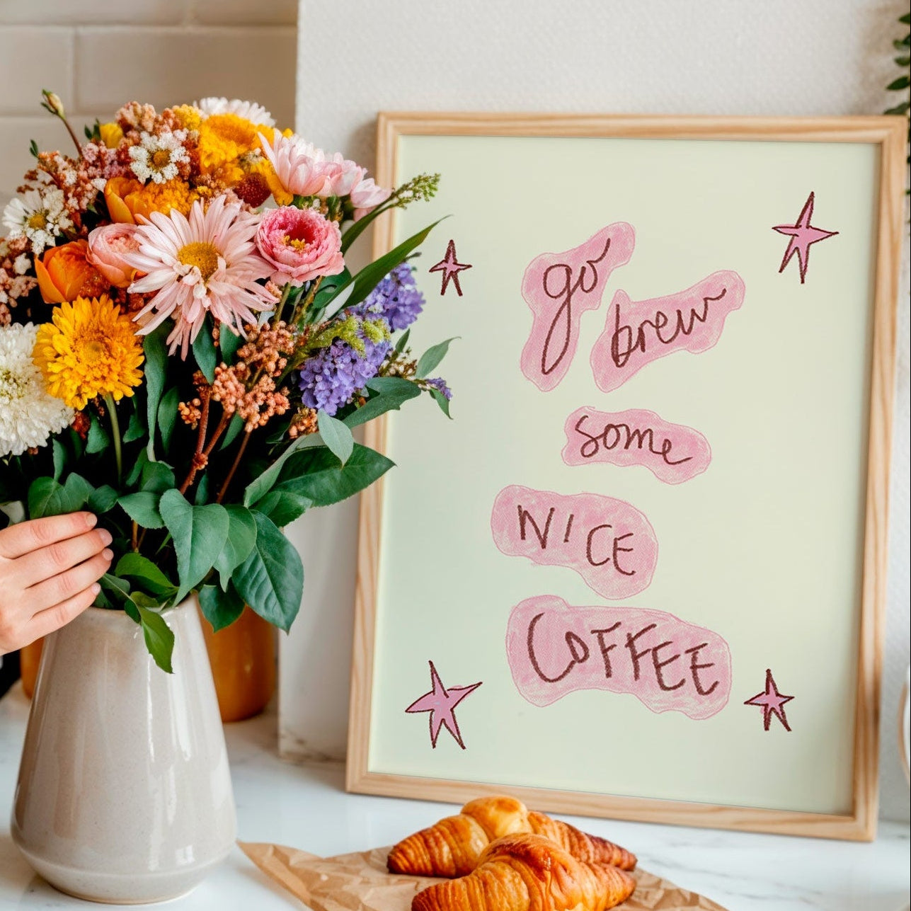 Person arranging flowers in a vase with a framed print that says 'go brew some nice coffee' and pastries on a table.