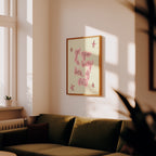 A framed art print with pink handwritten text and star illustrations on a cream background, hanging on a warm beige wall above a dark olive green sofa in a sunlit living room.