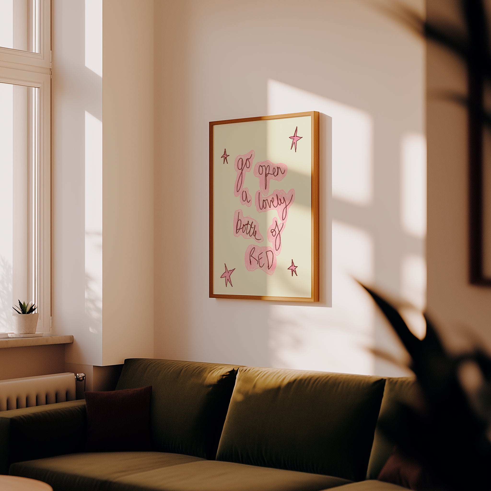 A framed art print with pink handwritten text and star illustrations on a cream background, hanging on a warm beige wall above a dark olive green sofa in a sunlit living room.