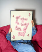 A person sitting on a textured magenta couch holds up a framed art print. The print features a cream background with pink hand-drawn bubble letters spelling out 'go open a lovely bottle of RED' and pink star accents, enclosed in a thin black frame.