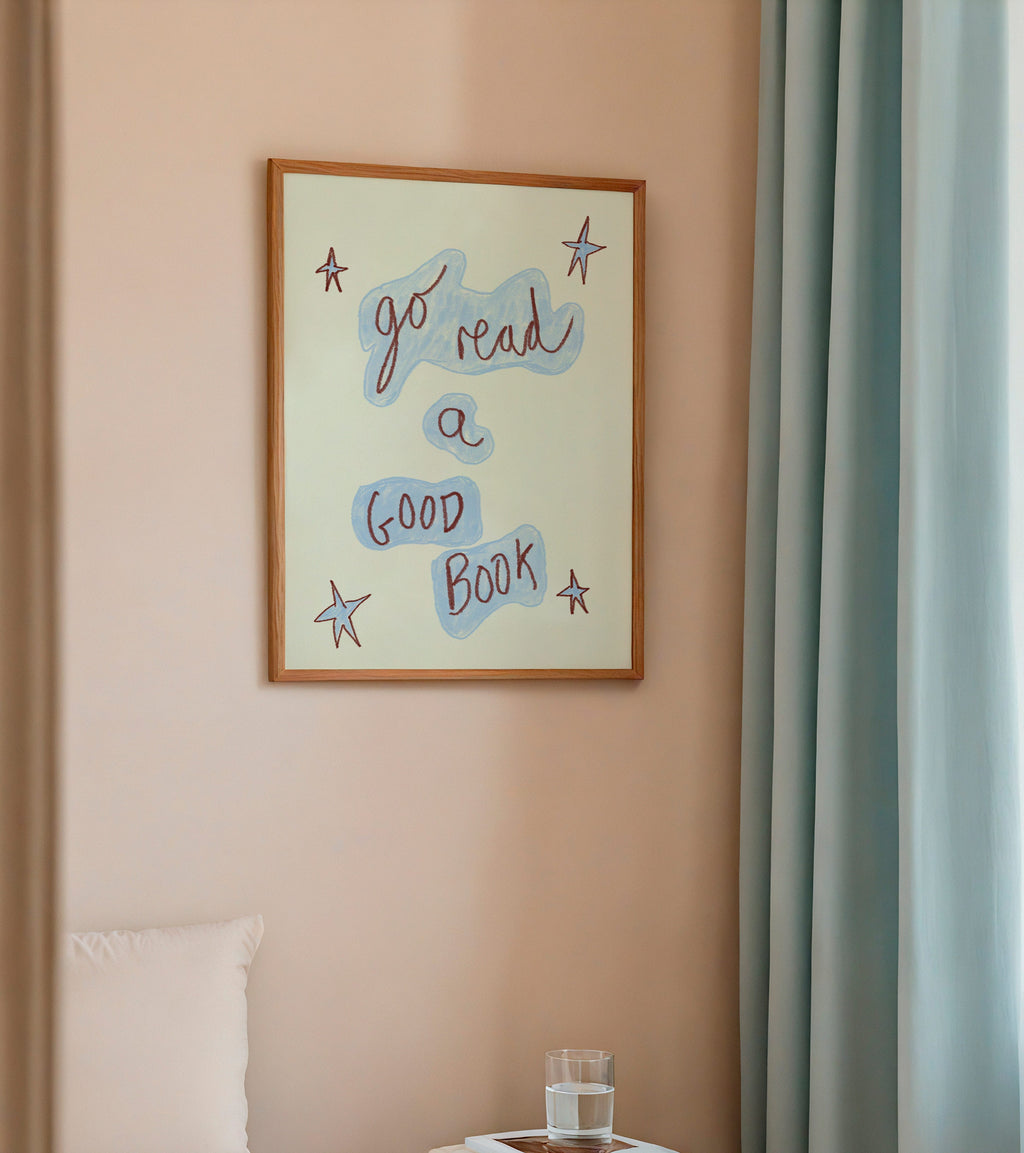 Framed artwork on a wall above a bed with a small table and books.