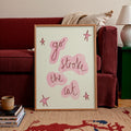 A framed art print leaning against a red sofa in a living room setting. The print features the hand-lettered phrase 'go stroke the cat' in dark brown script over pink abstract shapes, accented with pink star drawings on a cream background, enclosed in a light wood frame.