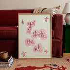 A framed art print leaning against a red sofa in a living room setting. The print features the hand-lettered phrase 'go stroke the cat' in dark brown script over pink abstract shapes, accented with pink star drawings on a cream background, enclosed in a light wood frame.