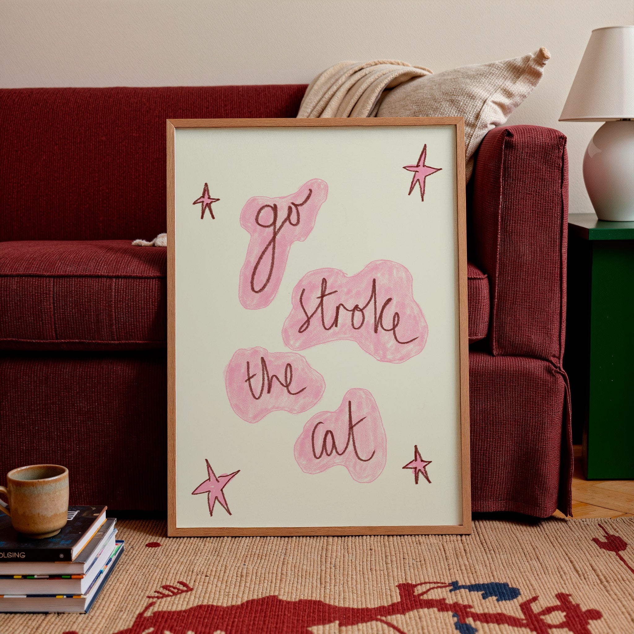 A framed art print leaning against a red sofa in a living room setting. The print features the hand-lettered phrase 'go stroke the cat' in dark brown script over pink abstract shapes, accented with pink star drawings on a cream background, enclosed in a light wood frame.