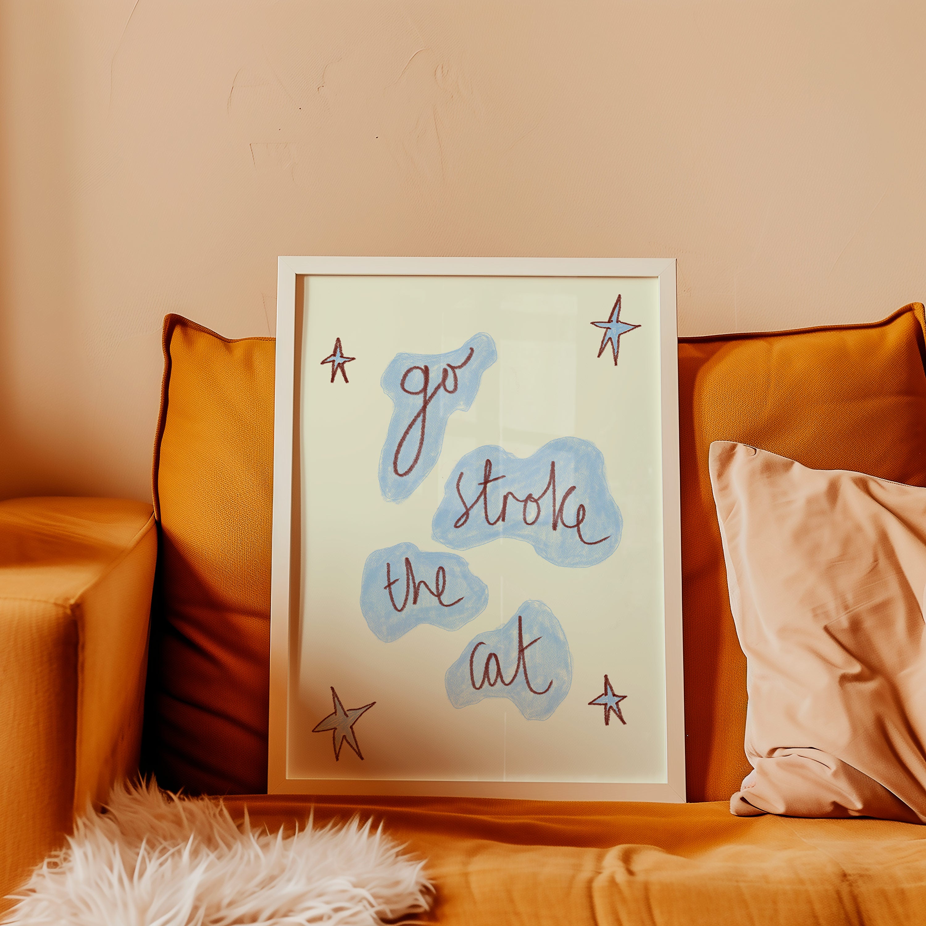 A framed art print with the phrase 'go stroke the cat' in cursive over light blue cloud shapes, styled on a mustard yellow sofa with pillows.