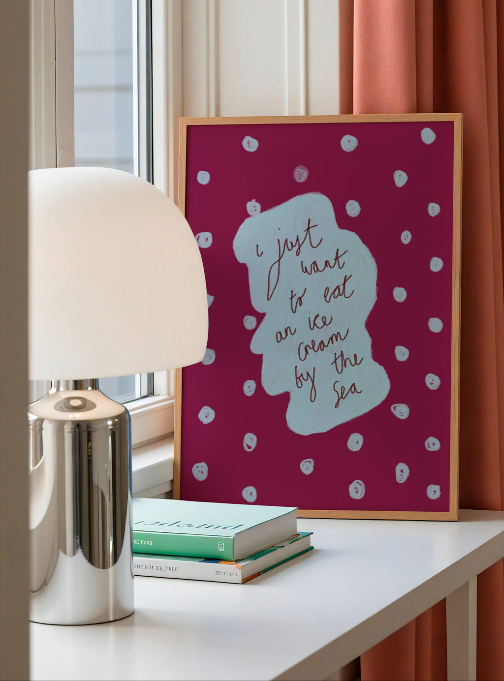 Framed artwork with text on a table next to a lamp and books, with a window and pink curtain in the background.