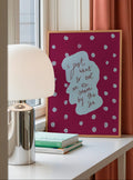 Framed artwork with text on a table next to a lamp and books, with a window and pink curtain in the background.
