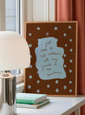 Framed artwork with text on a table next to a lamp and books, with pink curtains in the background.