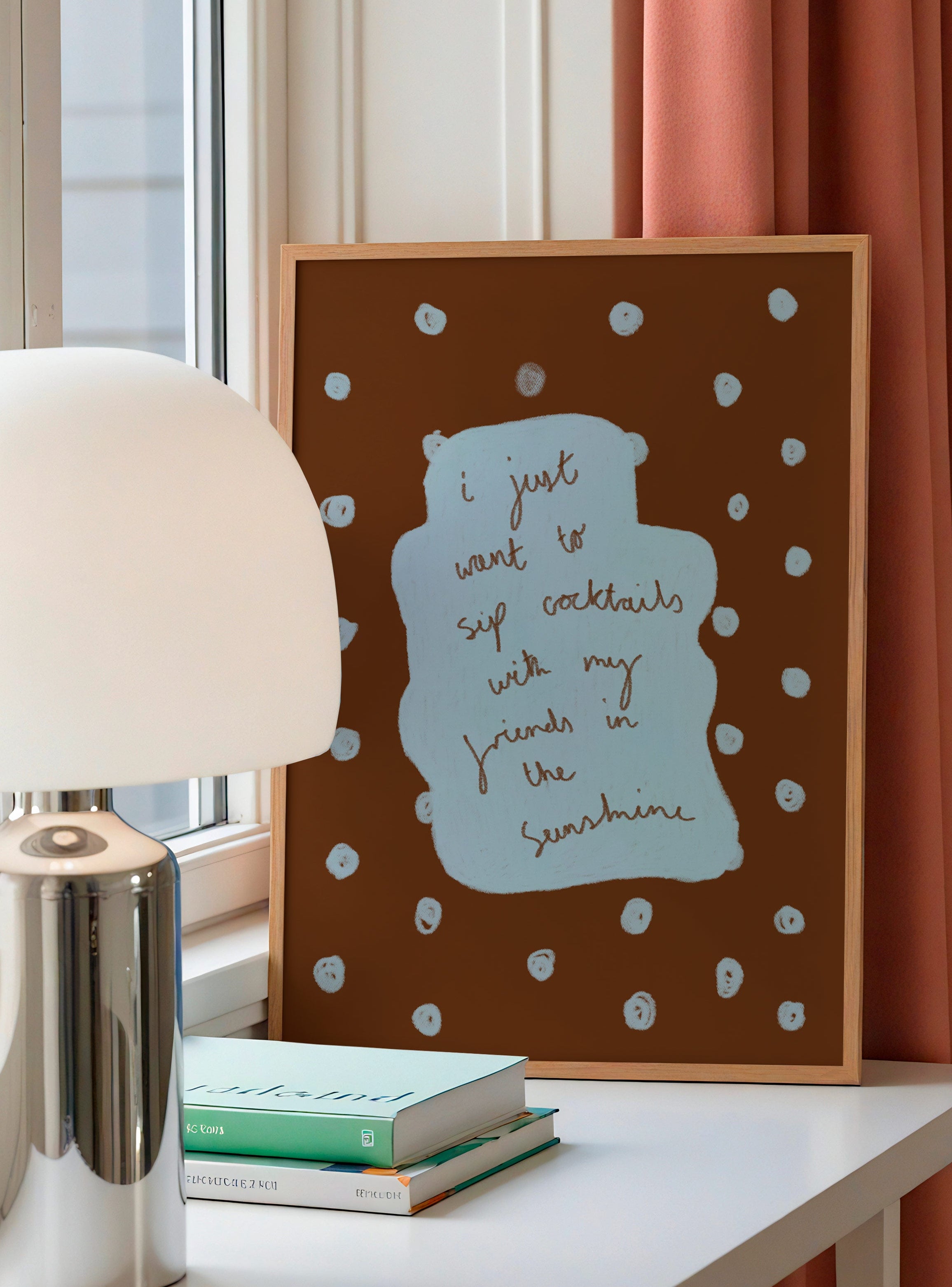 Framed artwork with text on a table next to a lamp and books, with pink curtains in the background.