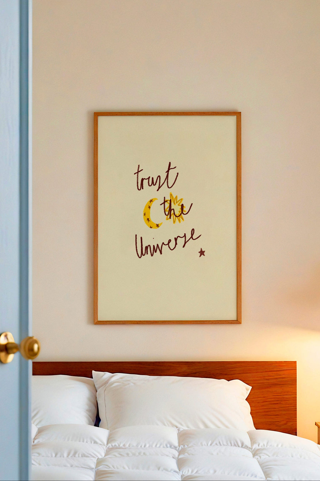 Bedroom with white bedding, wooden headboard, and framed artwork on wall with words 'trust the universe'.