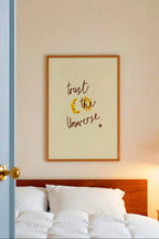 Bedroom with white bedding, wooden headboard, and framed artwork on wall with words 'trust the universe'.