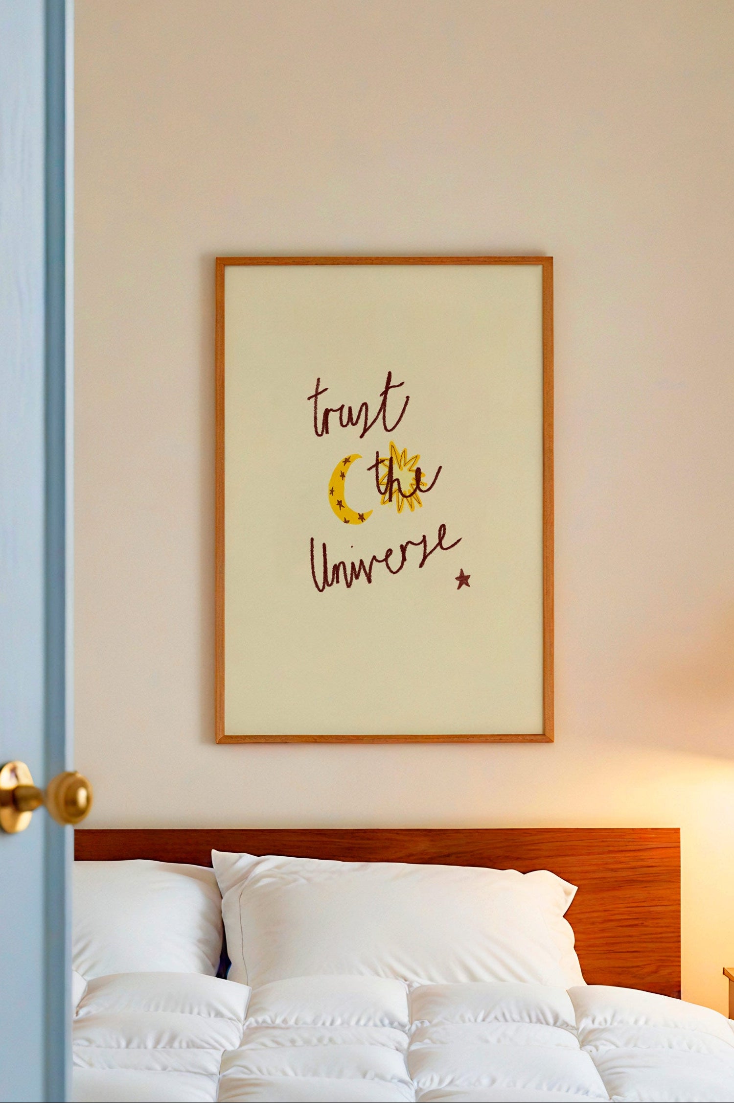 Bedroom with white bedding, wooden headboard, and framed artwork on wall with words 'trust the universe'.