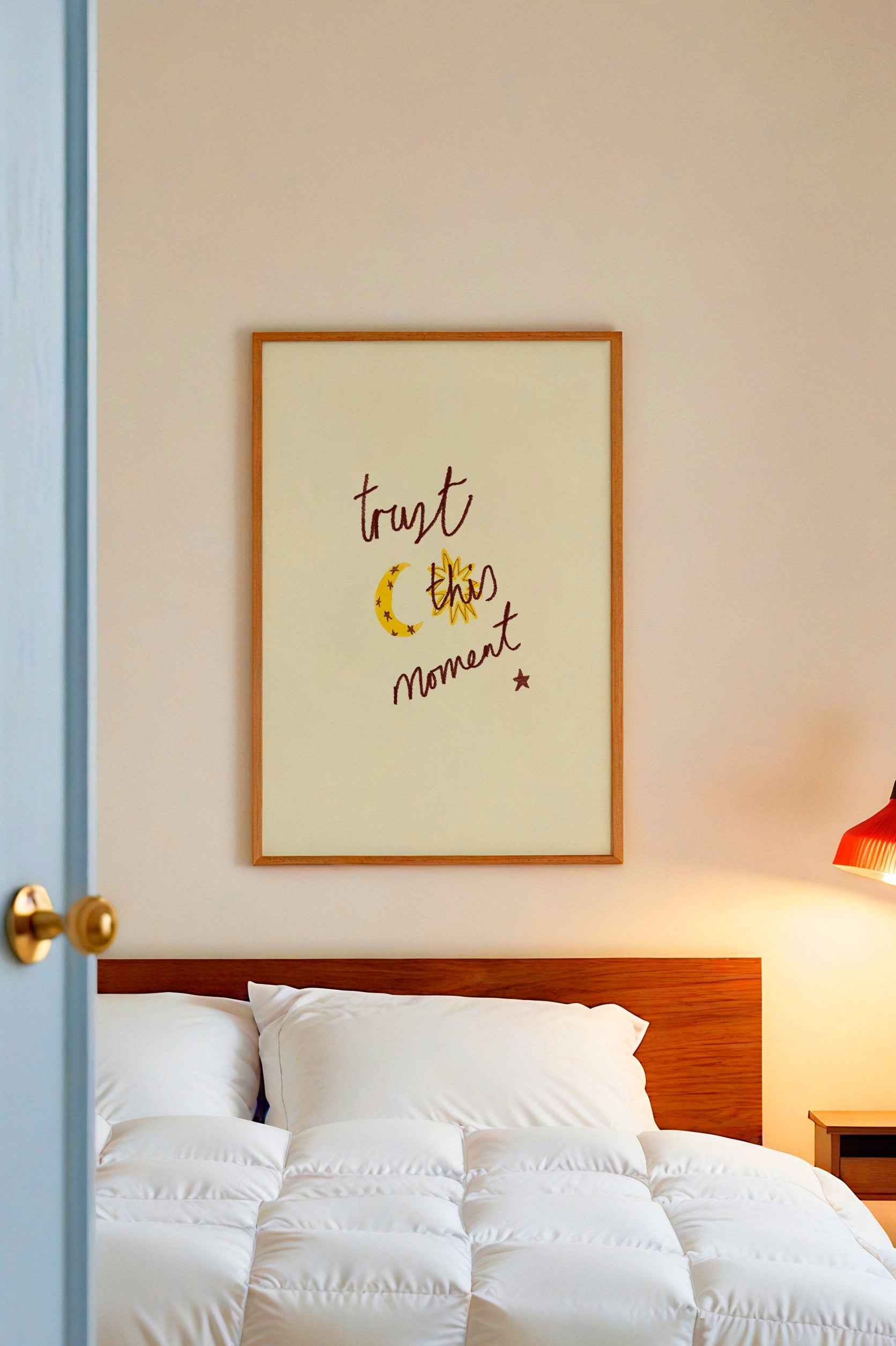Bedroom with white bedding, wooden headboard, and framed artwork on wall which says trust this moment.