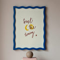 Framed artwork with motivational quote which reads 'trust the timing' on a wall above books