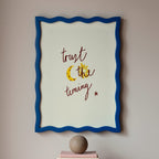 Framed artwork with motivational quote which reads 'trust the timing' on a wall above books