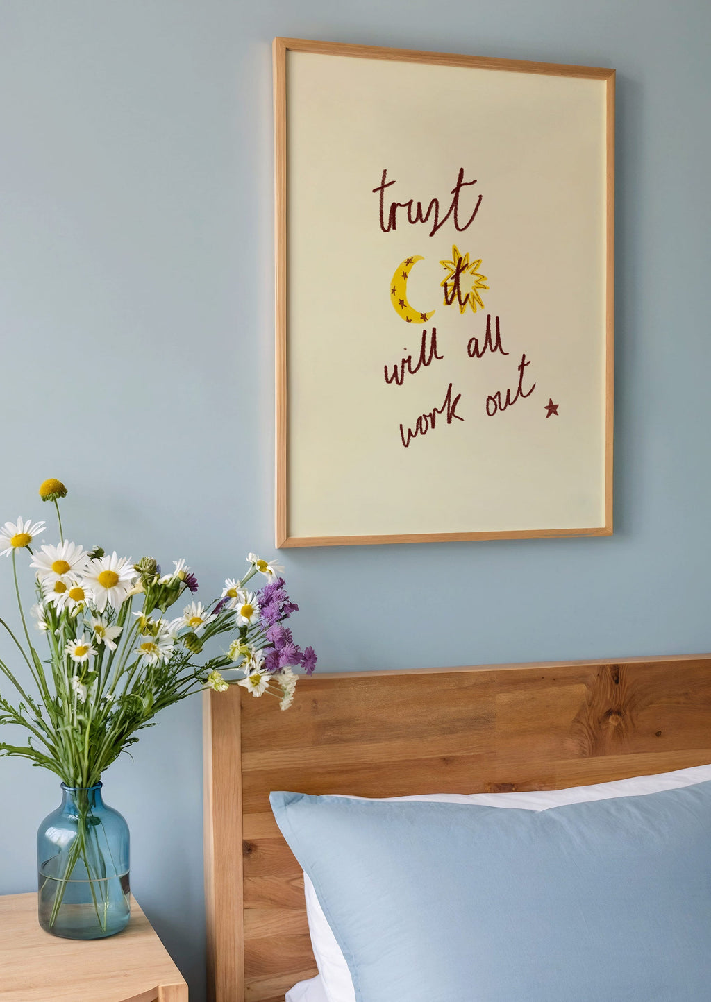 Bedroom with wooden headboard, blue pillows, and a vase of flowers on a nightstand. Framed print with 'trust it ill all work out