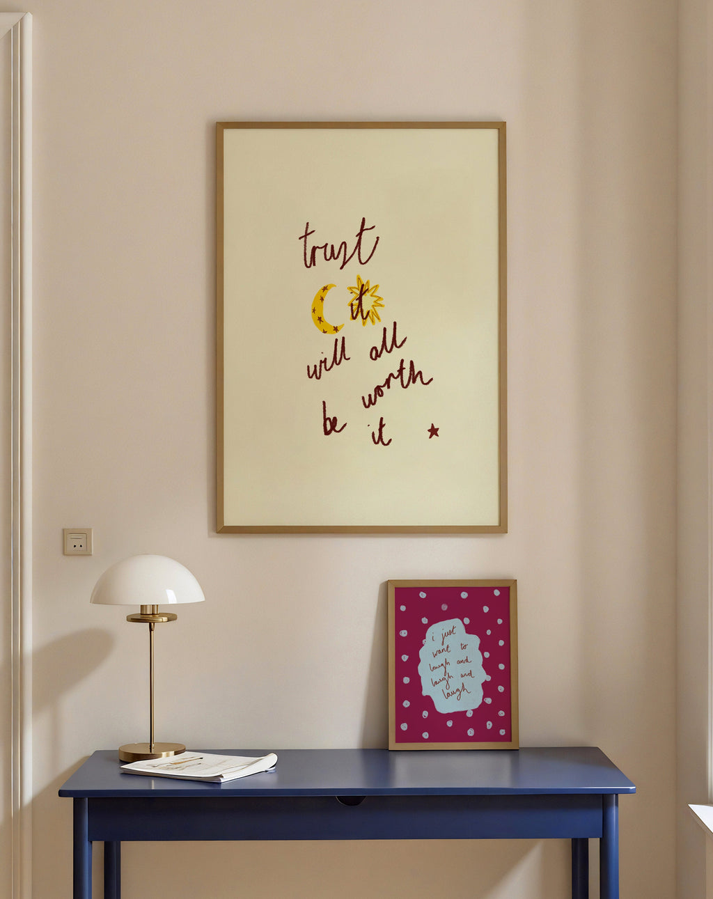 Framed artwork with phrase 'trust it will all be worth it' on a wall above a blue table with a lamp and book.